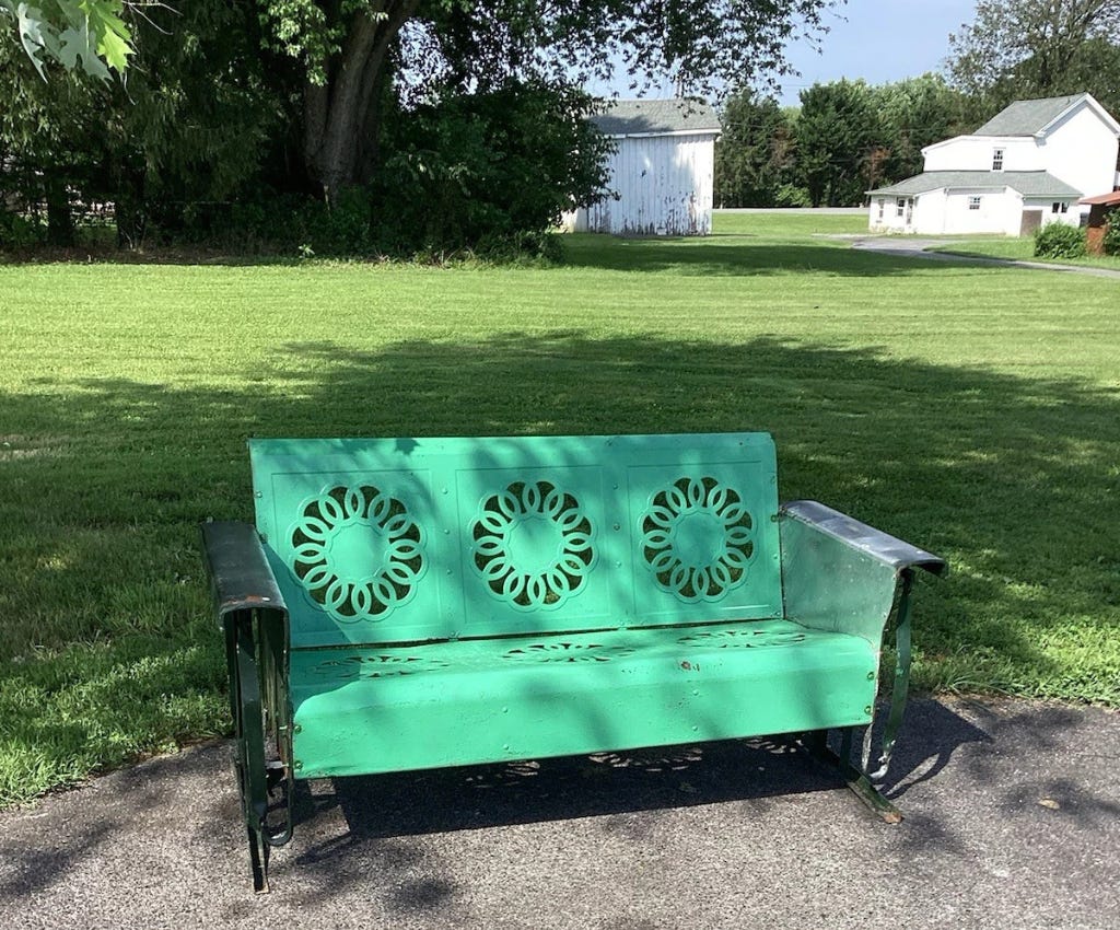 Bright mint green metal glider with three flower-like cut-out designs across the backrest and the seat. The old fashioned glider is sitting under the shade of an unseen tree on an asphalt drive surrounded by green grass. In the upper right corner is the backside of a large white clapboard well-kept farm house and garage. To the left of the garage is the back and side of a large white shed in need of painting.  The shed is in the shadow of a large tree and green bushes that extend to the left across the rest of the photo.