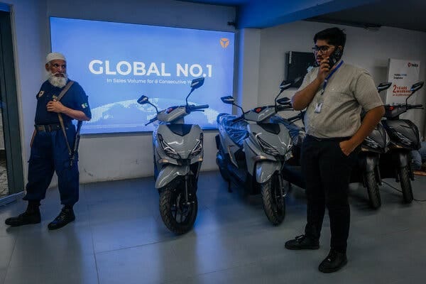 A person stands in front of a row of shiny gray motorbikes, speaking on a cellphone, while a security guard with a gun slung over his shoulder looks on.