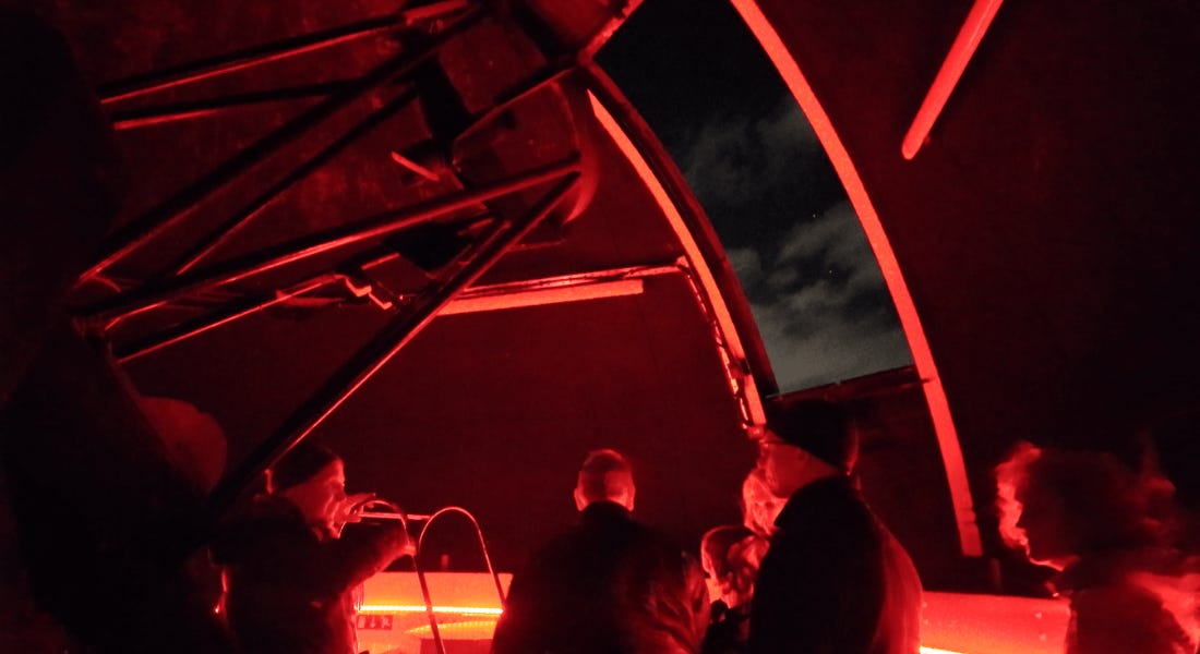 An observatory with dark red lights, a night sky panel, and people around