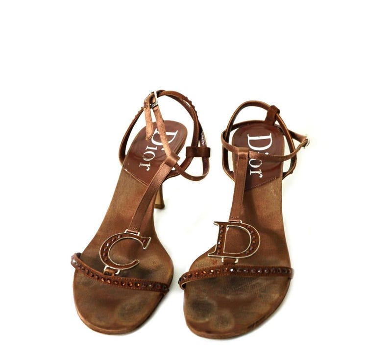 May include: A pair of brown leather heeled sandals with a t-strap design. The sandals have a gold metal 'CD' logo on the front strap and the word 'Dior' printed on the insole.
