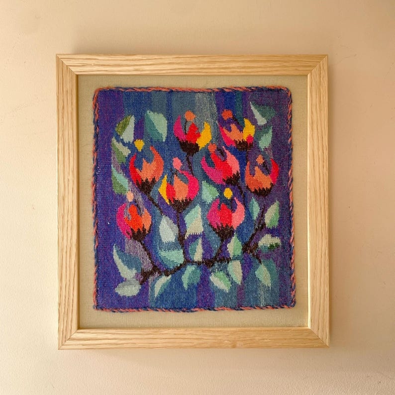 May include: Framed textile art featuring a floral design. The artwork showcases vibrant red, pink, and yellow flowers with green leaves against a blue background. The piece is encased in a light-colored wooden frame, suitable for wall decor.