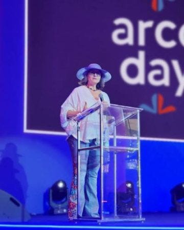 #descrevemospravocê  Em um palco iluminado com luzes azuis, uma mulher está em pé atrás de um púlpito transparente, segurando um microfone. Ela veste uma blusa branca com mangas largas, calça jeans bordada e um grande chapéu azul com abas largas e enfeites nas laterais. Atrás dela, vê-se parte de uma grande tela com o texto “arco day” e pequenos ícones coloridos em forma de corações estilizados. O fundo da tela é escuro com moldura clara, contrastando com o ambiente predominantemente azul. Luzes de palco estão posicionadas na parte inferior da imagem, apontadas para cima. A sombra da mulher é projetada na parede à esquerda, reforçando a iluminação intensa do local. A composição da imagem transmite um clima de evento ou apresentação formal com toques de estilo pessoal e descontração.