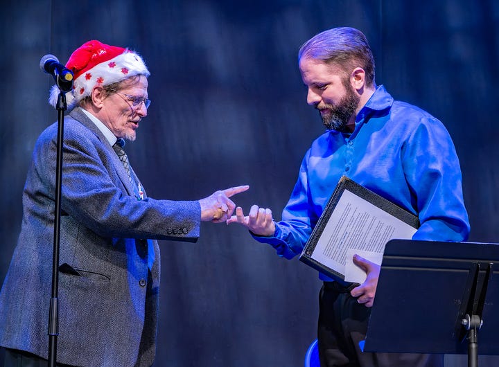 Two men stand on a stage under blue lighting. On the left, an older man in a gray blazer, tie, glasses, and a Santa hat gestures with a pointed finger. On the right, a bearded man in a bright blue shirt holds a folder with printed pages and looks at his hand as if responding. A microphone on a stand is beside them, and a music stand is in the foreground, suggesting a performance or reading. Also, A festive scene in a building lobby: four adults stand behind and around a holidayâdecorated information table. In front, a man in a Santa hat and a woman in a red blouse shake hands and smile. Behind them, two others (one wearing a Santa hat, another with a long white beard and dog tags) stand by the table, which is covered with a blue, starâspangled cloth, gold runner, ornaments, and brochures. A sandwichâboard sign on the left reads âTallahassee Veterans Legal Clinicâ with details for Thursdays at American Legion Post 13, 229 Lake Ella Drive, Tallahassee, Florida. The background shows a wooden door marked âStairs,â glassâblock windows, and a wallâmounted TV.