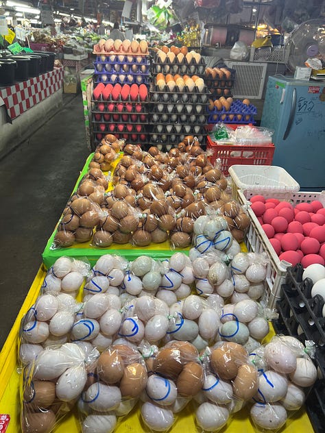Outdoor farmers market in Chiang Mai with multicolored eggs, rows of produce, meats, and crickets.