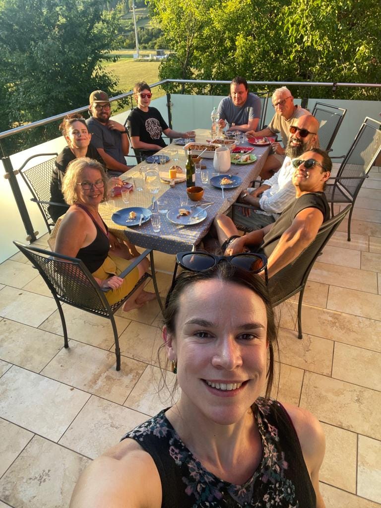 On a sun-dappled balcony, a family gathers for dinner. A white- and gray-haired couple, their adult children with their partners, and a nephew with his wife. In the foreground is a woman creating the shot, a selfie. On a sun-dappled balcony, a family gathers for dinner. A white- and gray-haired couple, their adult children with their partners, and a nephew with his wife. In the foreground is a woman creating the shot, a selfie.