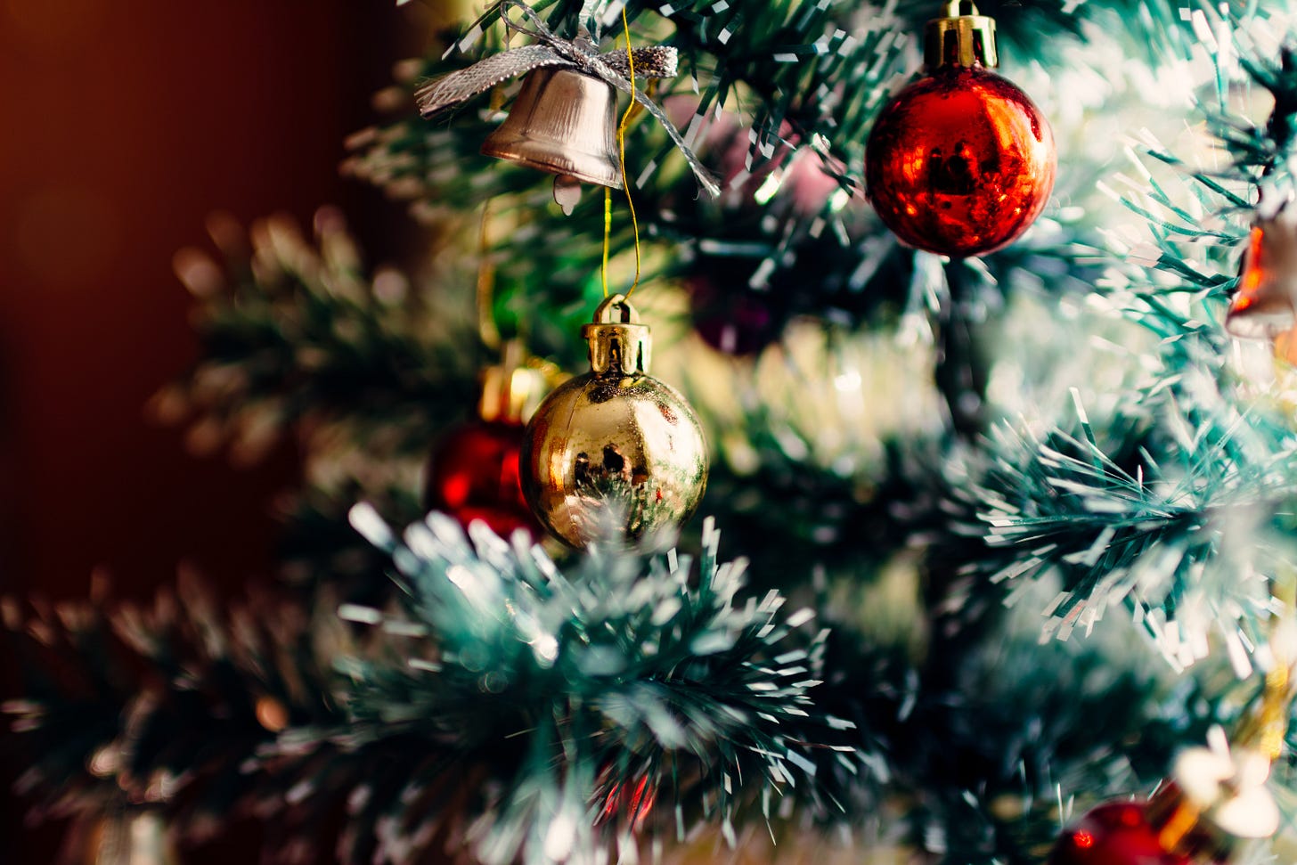 Foto em close de uma bolinha dourada pendurada em uma árvore de natal de plástico. logo acima dela está um sino também dourado.as folhas são verdes com as pontas brancas, no fundo desfocado estão outras bolinhas vermelhas.