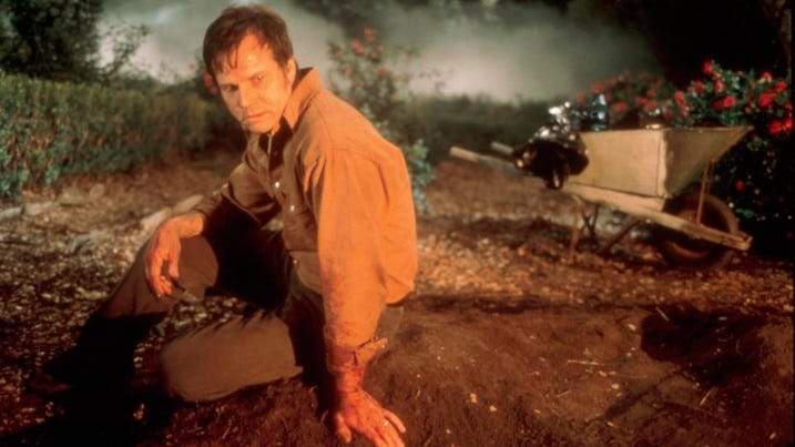 Bloodied and exhausted, Bill Paxton sits in a dirt patch near a wheelbarrow and rose bushes, staring forward with intensity.