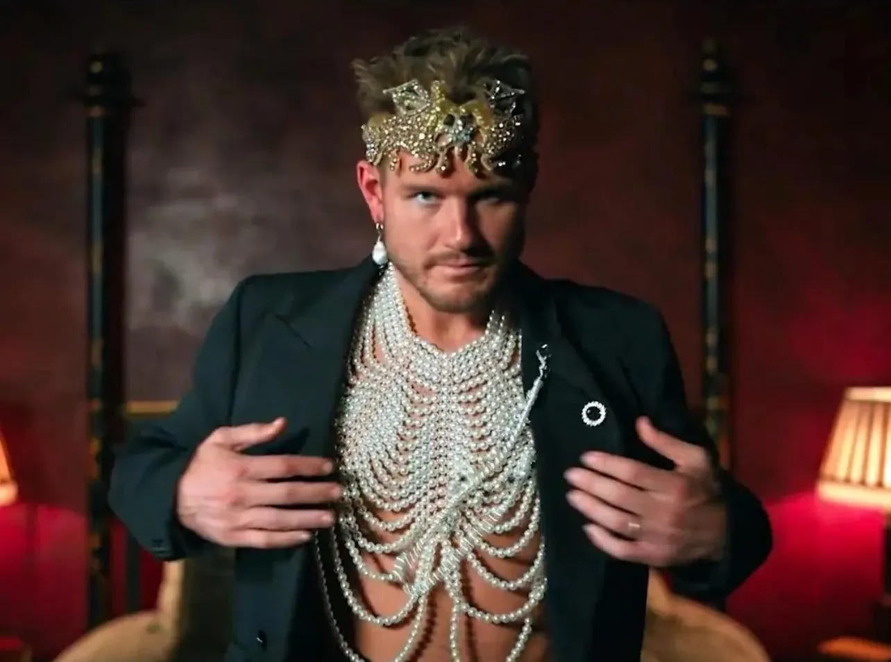 Colton in a dimly lit confessional, blazer open to reveal a draped pearl body harness over bare chest, gold crown with centaur designs, pearl drop earring.
