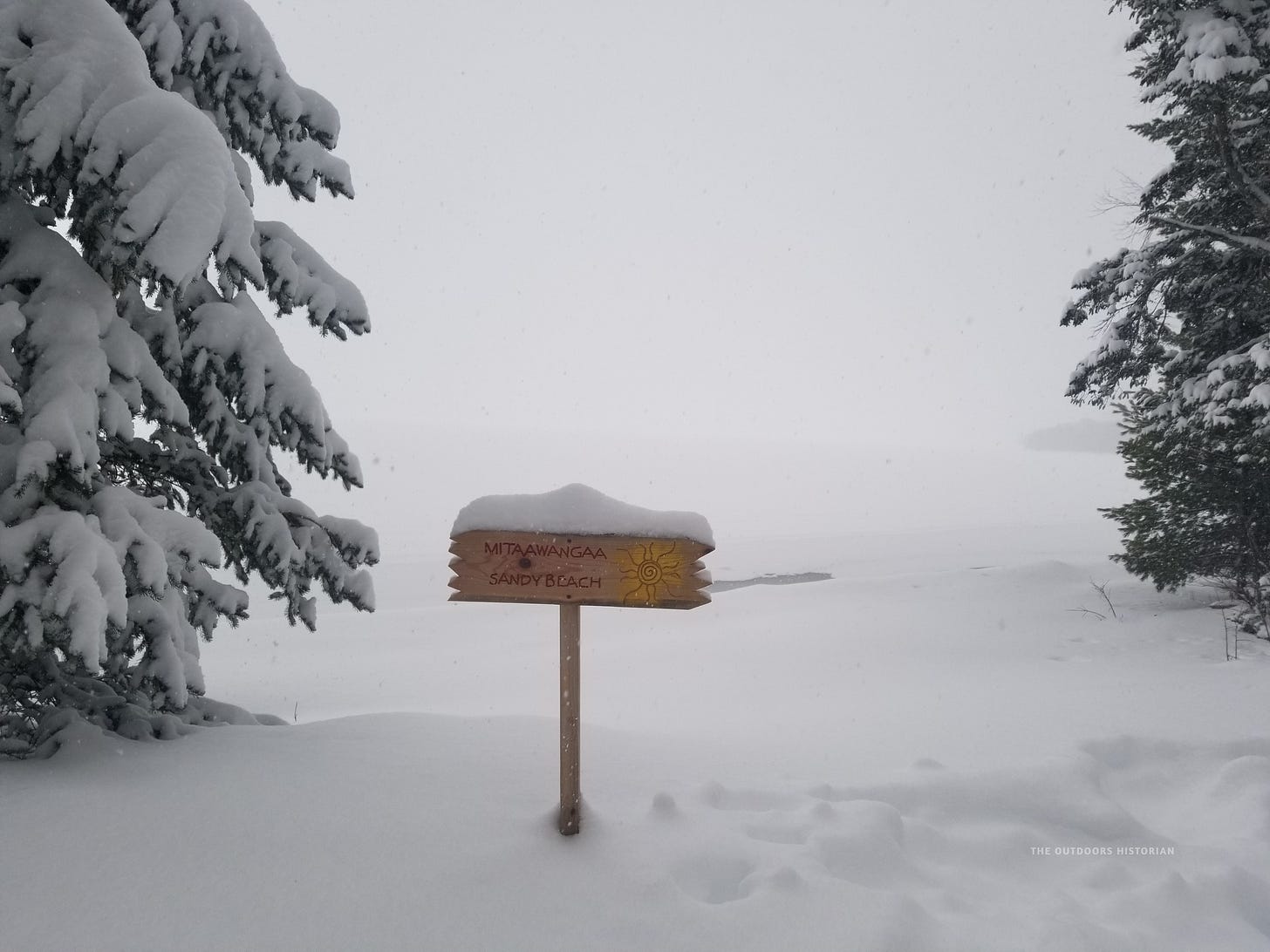 A landscape photograph of the Frog Bay shoreline in winter, covered in a deep blanket of white snow against a frozen, icy Lake Superior. In the frame, a wooden park sign clearly displays the Anishinaabemowin word "Mitaawanga," translating to "sandy beach."