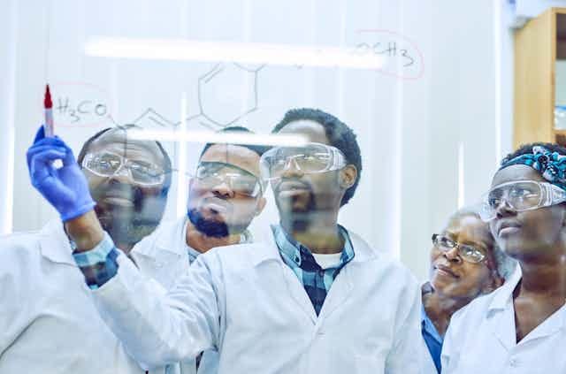 Five people in white coats and protective eyewear look at a clear wall on which one is writing a formula