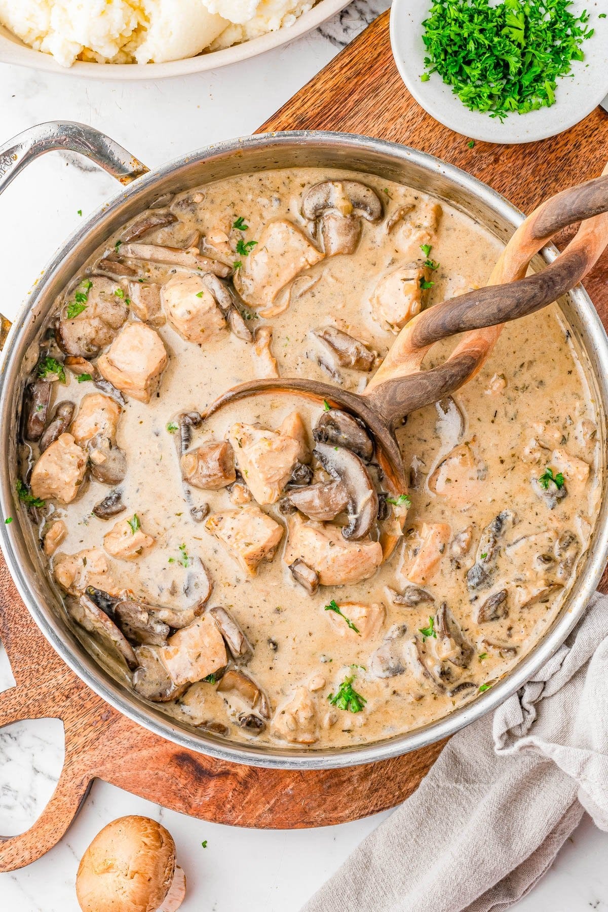 A skillet filled with creamy mushroom and chicken stew, garnished with chopped parsley, with a wooden spoon and ladle resting in the pan. A skillet filled with creamy mushroom and chicken stew, garnished with chopped parsley, with a wooden spoon and ladle resting in the pan.