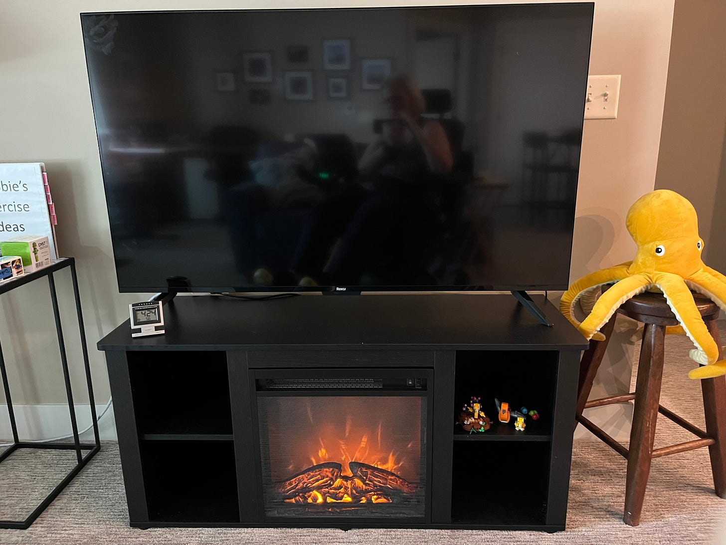 A large TV is supported by a black entertainment stand with a glowing fireplace in the middle. On one side is an antique stool with a yellow octopus on top and the other side is a stand holding weights and other exercise equipment.
