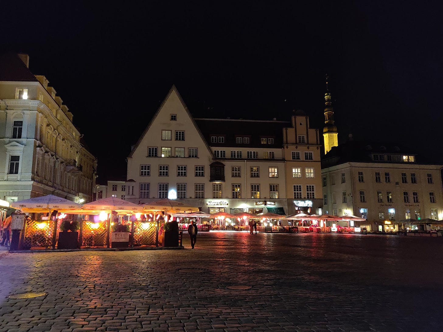 Tallinn's old town square at night, lit restaurant umbrellas and a person walking across wet cobblestones.