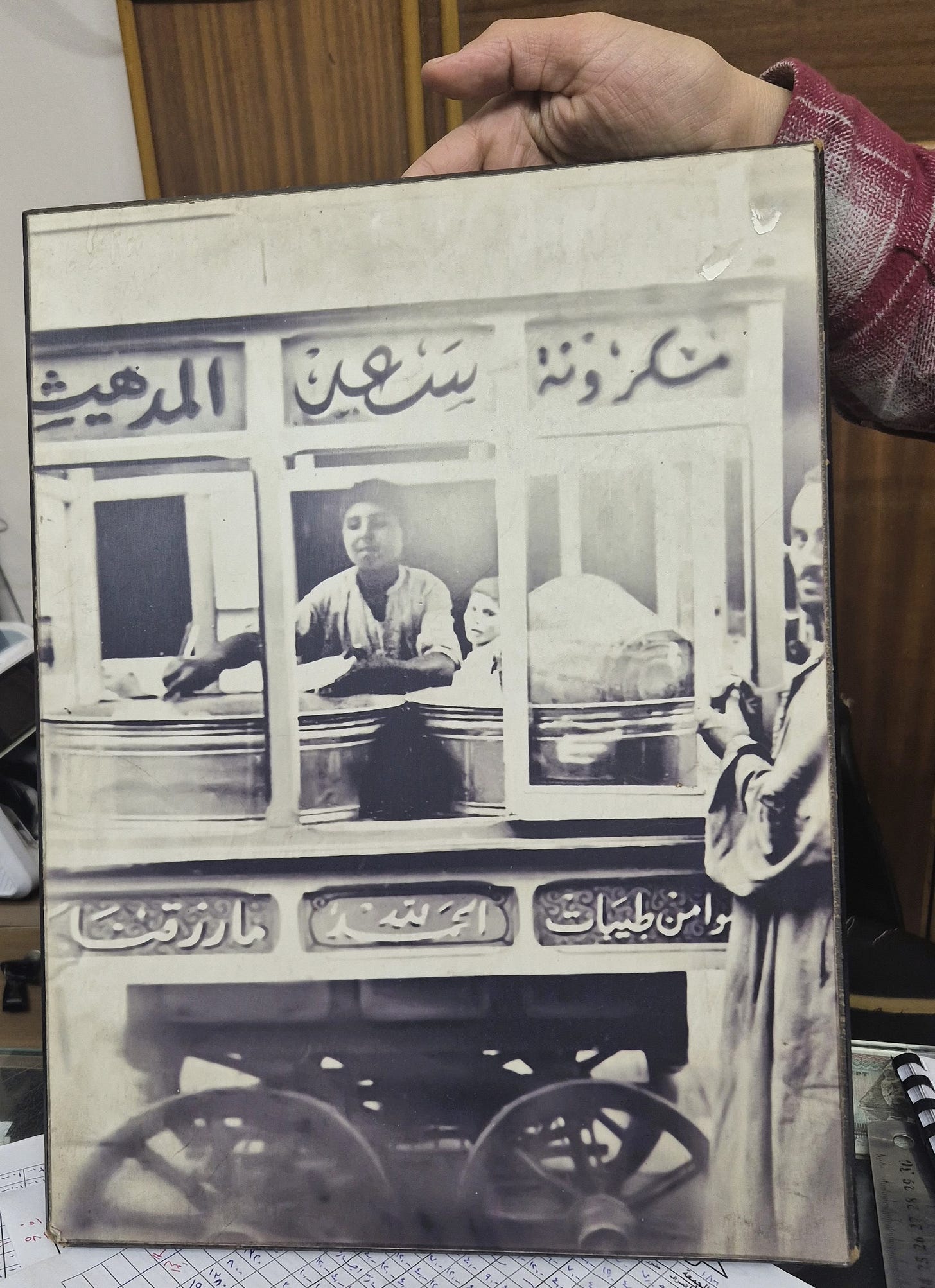 a hand holding up a framed historical photo in black and white of a street food cart with a boy scooping from two large metal rounds