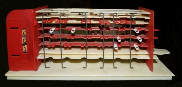 Photo of an original Digi-Comp I plastic, mechanical computer, ca. 1963. It is made of layers of red & black plastic, with wires and small plastic tubes that manipulate gears to literally crank out 1s & 0s. A small window to the left of the machine displays 3 wheels with ones or zeros to show answers from 0 to 7.