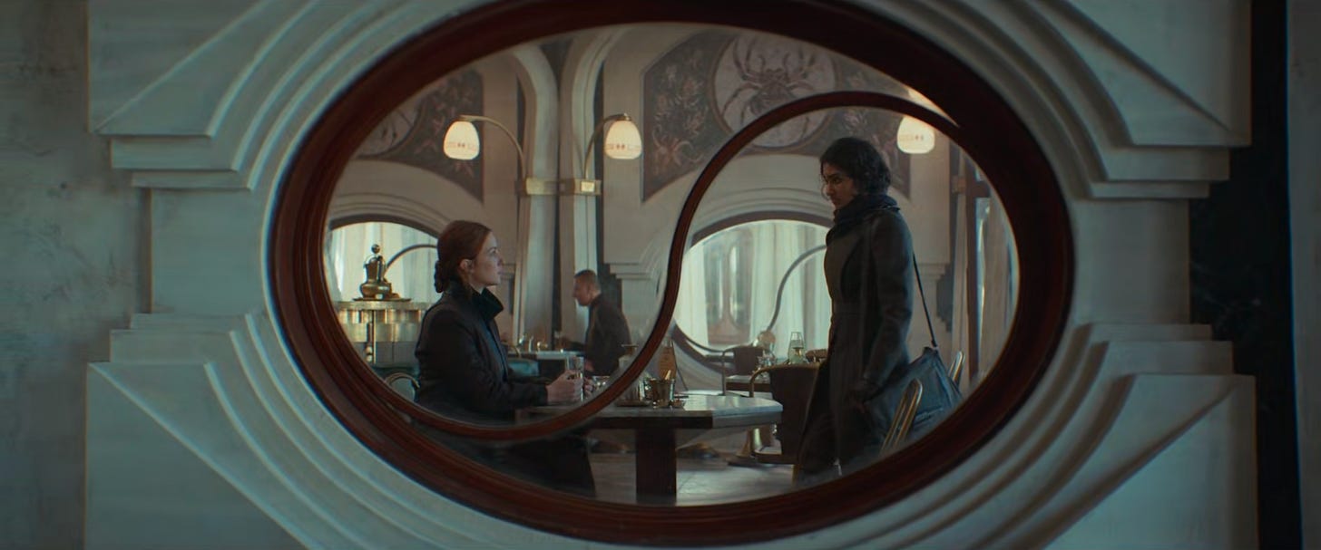 a medium shot of a cafe window; the window is oval-shaped, with an unusual middle frame piece which creates a yin-yang outline.  On the left, outlined in the 'bottom' part of the yin-yang symbol, a woman with red hair is sitting.  On the right, outlined in the 'top' part of the yin-yang symbol, a woman with black hair is standing.