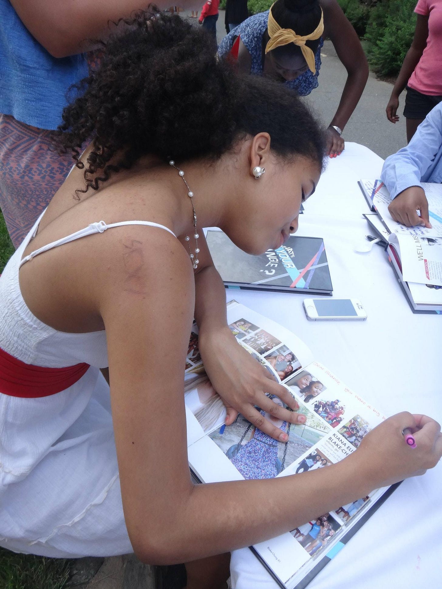 Kiana in a white dress signing her page in the yearbook for a friend.