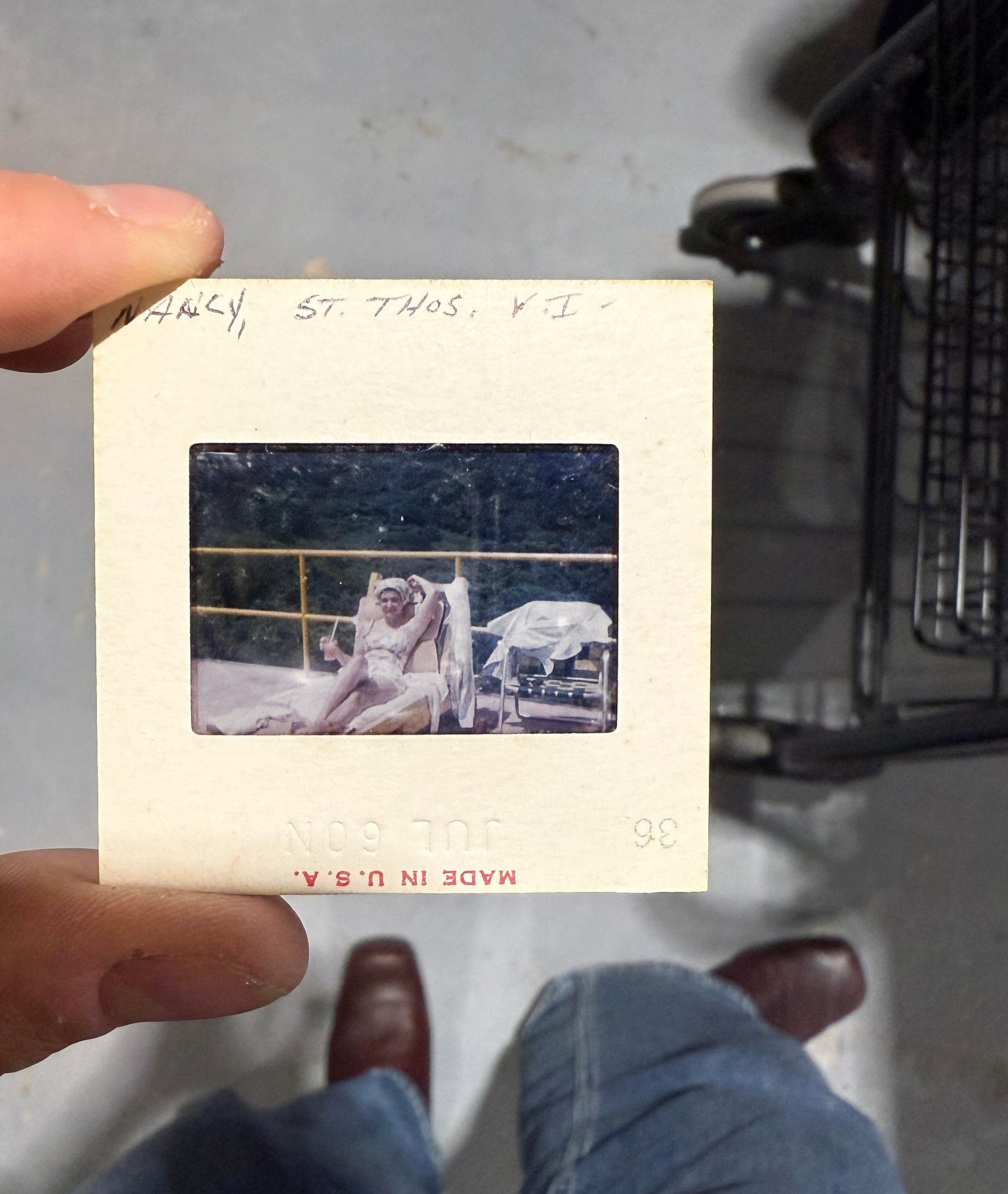 A hand holding a Kodak slide up to fluorescent light, showing a vintage photo of a woman in white sitting on a boat deck with blue water in the background A hand holding a Kodak slide up to fluorescent light, showing a vintage photo of a woman in white sitting on a boat deck with blue water in the background
