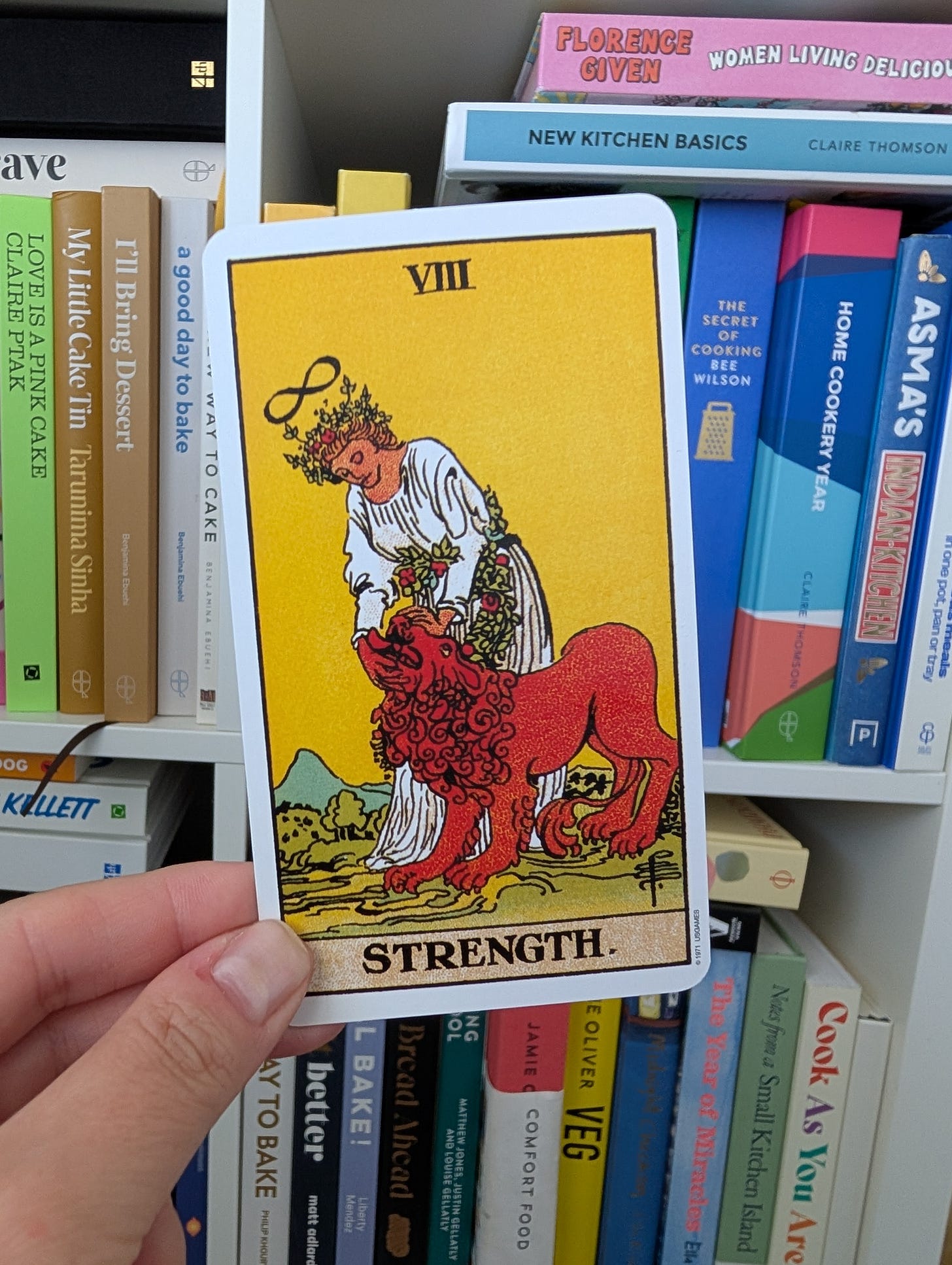 A hand holds up a tarot card labelled "VIII STRENGTH" in front of a bookshelf filled with colourful cookbooks. The card depicts a woman in a white robe gently taming a red lion, symbolising inner strength, compassion, and resilience. An infinity symbol floats above her head. Book titles visible on the shelf include "I'll Bring Dessert", "New Kitchen Basics", "The Secret of Cooking", and "Women Living Deliciously".