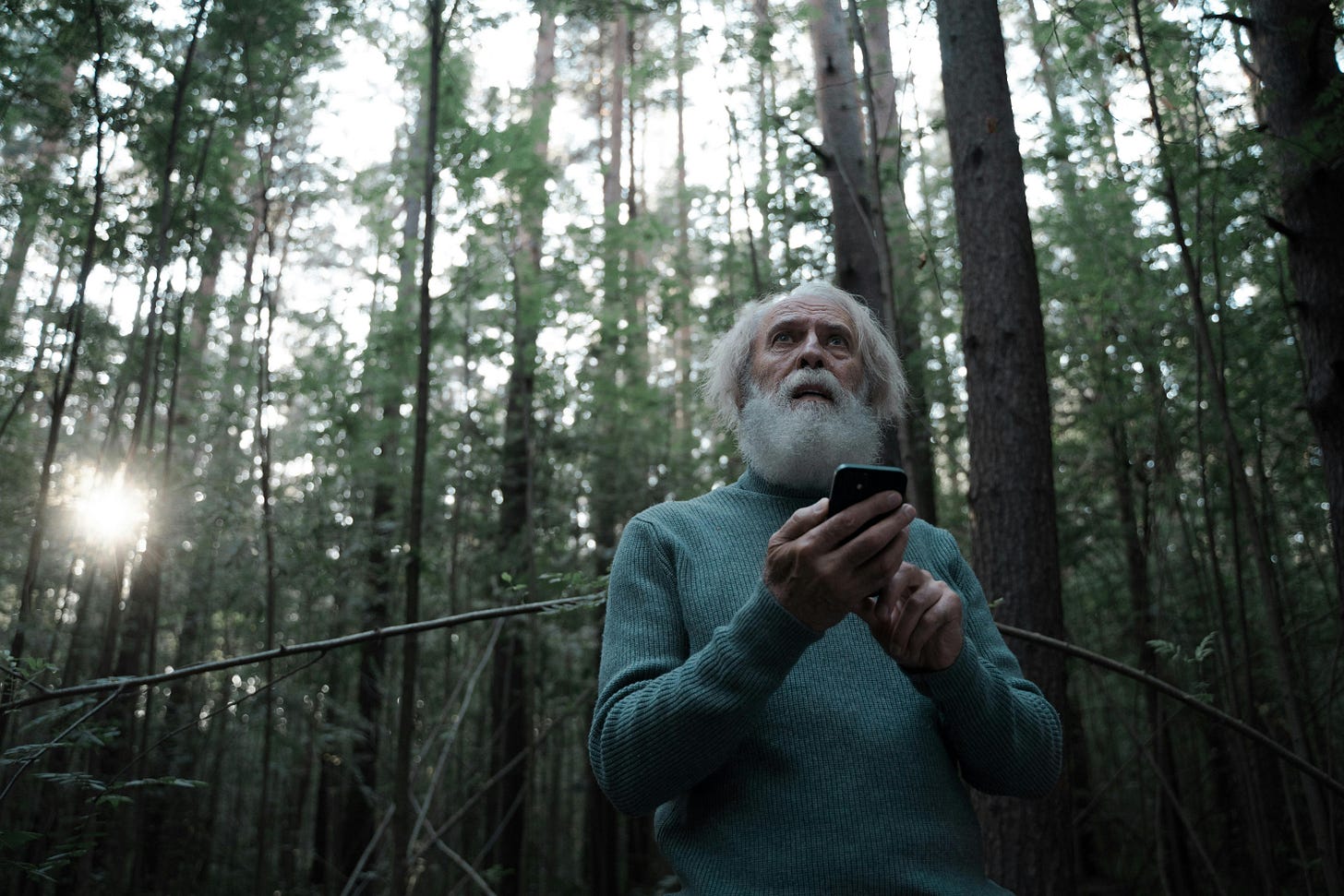 Um homem mais velho está em pé em meio a uma floresta densa. Ele tem cabelos longos e brancos, assim como uma barba cheia e branca, que lhe dá uma aparência sábia e serena. Está vestindo um suéter verde-azulado, que contrasta suavemente com o ambiente natural ao seu redor. A floresta é composta por árvores altas e retas, com folhagens verdes que filtram a luz do sol. Os raios solares penetram entre os galhos, criando um jogo de luz e sombra que dá à cena um ar tranquilo e um pouco misterioso. O homem segura um smartphone com uma das mãos e olha para cima, como se estivesse buscando sinal, observando algo nas copas das árvores ou simplesmente contemplando o céu. A presença do celular em meio à natureza cria um contraste interessante entre o mundo moderno e o ambiente selvagem, sugerindo temas como conexão, isolamento ou busca por orientação. Um homem mais velho está em pé em meio a uma floresta densa. Ele tem cabelos longos e brancos, assim como uma barba cheia e branca, que lhe dá uma aparência sábia e serena. Está vestindo um suéter verde-azulado, que contrasta suavemente com o ambiente natural ao seu redor. A floresta é composta por árvores altas e retas, com folhagens verdes que filtram a luz do sol. Os raios solares penetram entre os galhos, criando um jogo de luz e sombra que dá à cena um ar tranquilo e um pouco misterioso. O homem segura um smartphone com uma das mãos e olha para cima, como se estivesse buscando sinal, observando algo nas copas das árvores ou simplesmente contemplando o céu. A presença do celular em meio à natureza cria um contraste interessante entre o mundo moderno e o ambiente selvagem, sugerindo temas como conexão, isolamento ou busca por orientação.