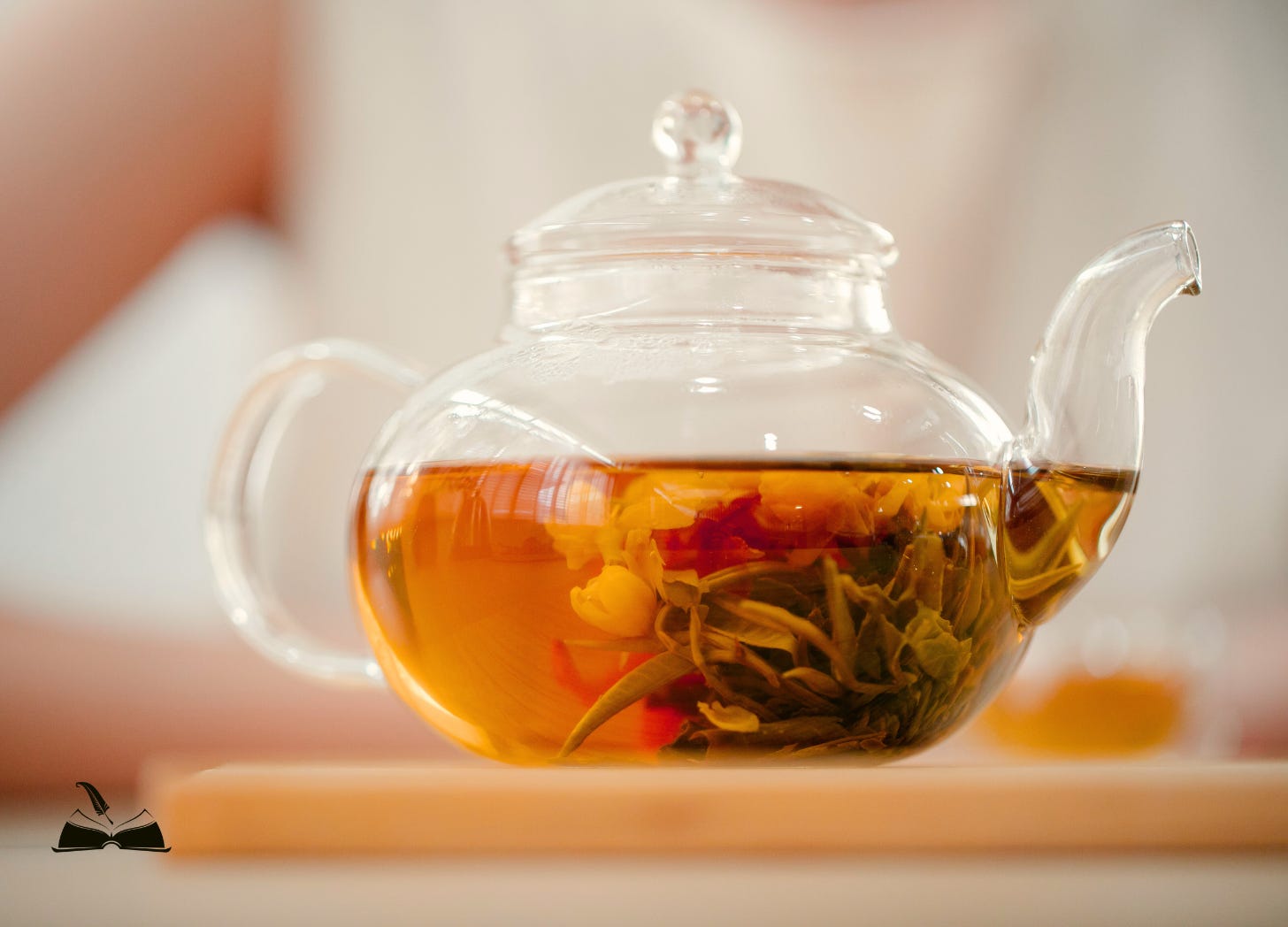 A close-up of a clear glass teapot filled with steeping loose-leaf tea and flower petals. The water is a warm amber color, and the teapot rests on a light wooden board.
