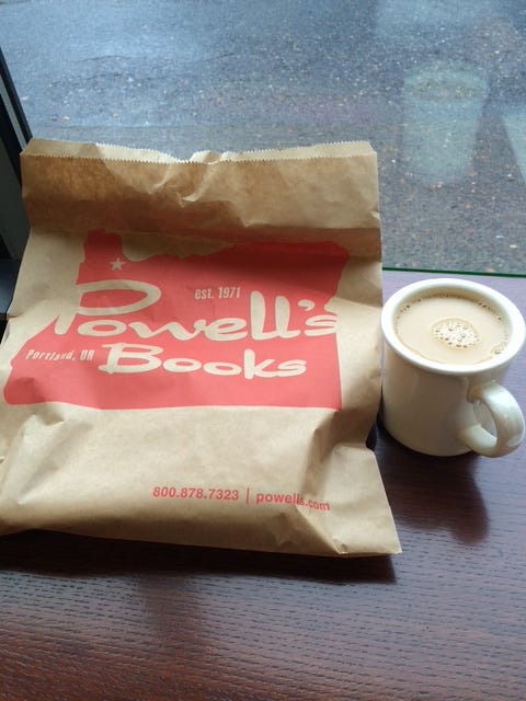 A paper bag has a design of the state of Oregon and the words Powell's Books. Next to it is a full mug of coffee.