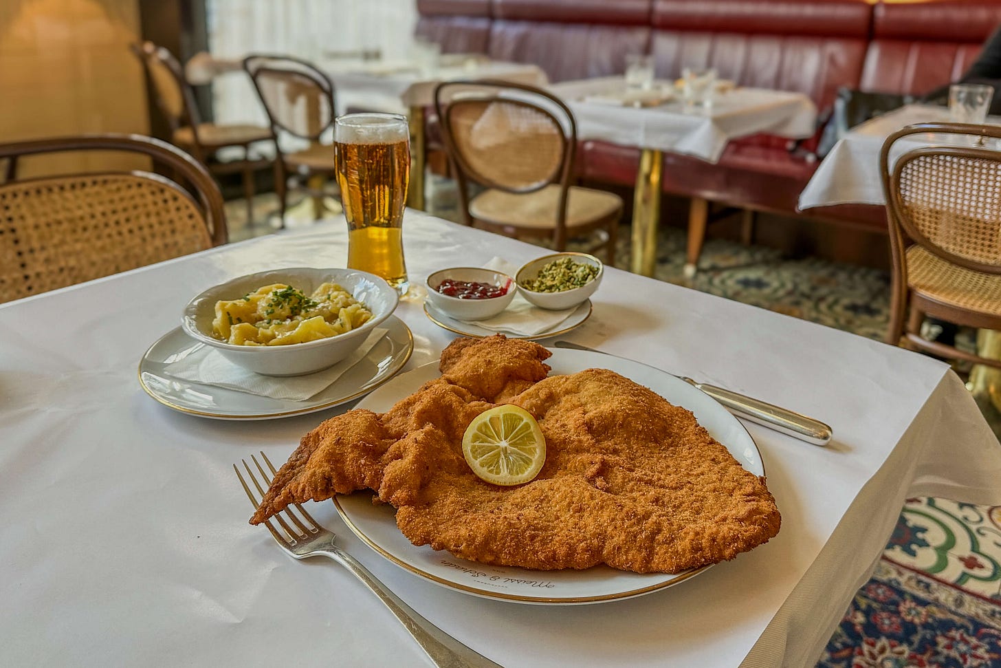Ein Wiener Schnitzel auf einem grossen Teller mit Zitronenscheibe in einem traditionellen Restaurant mit weissen Tischtüchern. Daneben stehen eine Schüssel Erdäpfelsalat, kleine Schalen mit Preiselbeeren und Petersilienbröseln sowie ein Glas Bier. Im Hintergrund sind Holzstühle und rote Polsterbänke zu sehen.