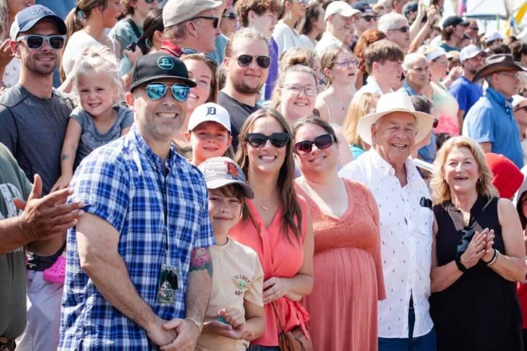 A large group of people, including adults and children, stand outdoors in sunny weather, smiling and clapping while facing the camera—clearly celebrating Connor’s big news.