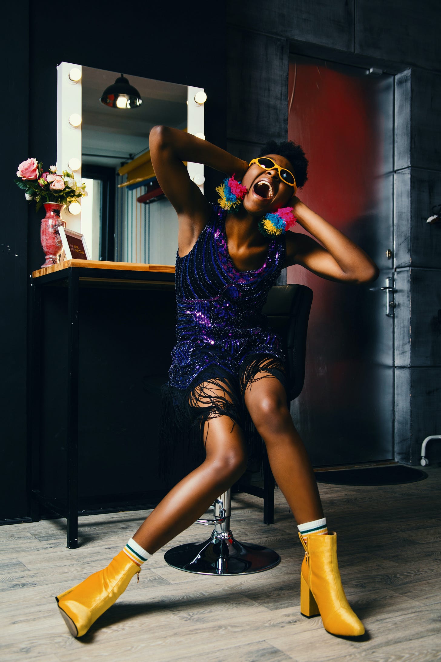A model covering her ears is wearing a shiny dress, bright yellow boots, sunglasses and pom-pom earrings.