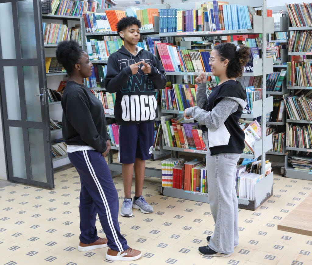 Três pessoas em pé em uma biblioteca, cercadas por estantes de livros coloridos, sendo que a pessoa do centro veste moletom preto com estampa branca, a pessoa à esquerda veste moletom preto com calça azul marinho com listras brancas e tênis marrom, e a pessoa à direita veste jaqueta preta com cinza e calça jeans clara, todas conversando entre si em um piso de cerâmica com padrão geométrico. Três pessoas em pé em uma biblioteca, cercadas por estantes de livros coloridos, sendo que a pessoa do centro veste moletom preto com estampa branca, a pessoa à esquerda veste moletom preto com calça azul marinho com listras brancas e tênis marrom, e a pessoa à direita veste jaqueta preta com cinza e calça jeans clara, todas conversando entre si em um piso de cerâmica com padrão geométrico.
