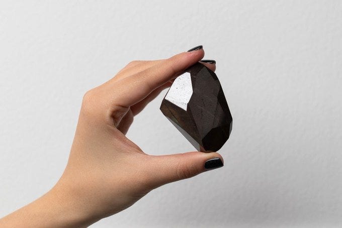 A hand with dark nail polish holds a large faceted black diamond against a plain grey background, referencing the Enigma black diamond purchased by Richard Heart for $4.3 million in cryptocurrency A hand with dark nail polish holds a large faceted black diamond against a plain grey background, referencing the Enigma black diamond purchased by Richard Heart for $4.3 million in cryptocurrency