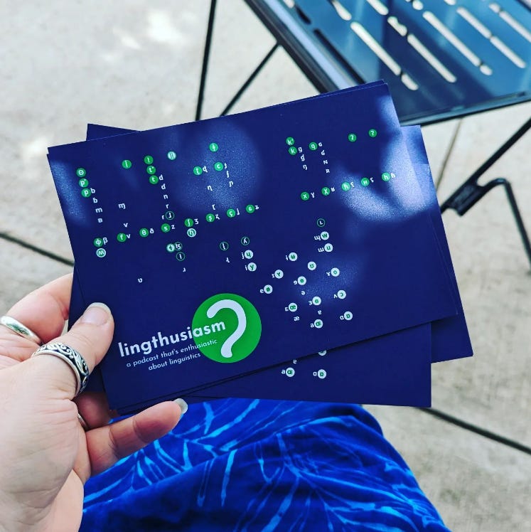 Left hand with rings holding blue cards with Lingthusiasm logo and green and white International Phonetic Alphabet. 