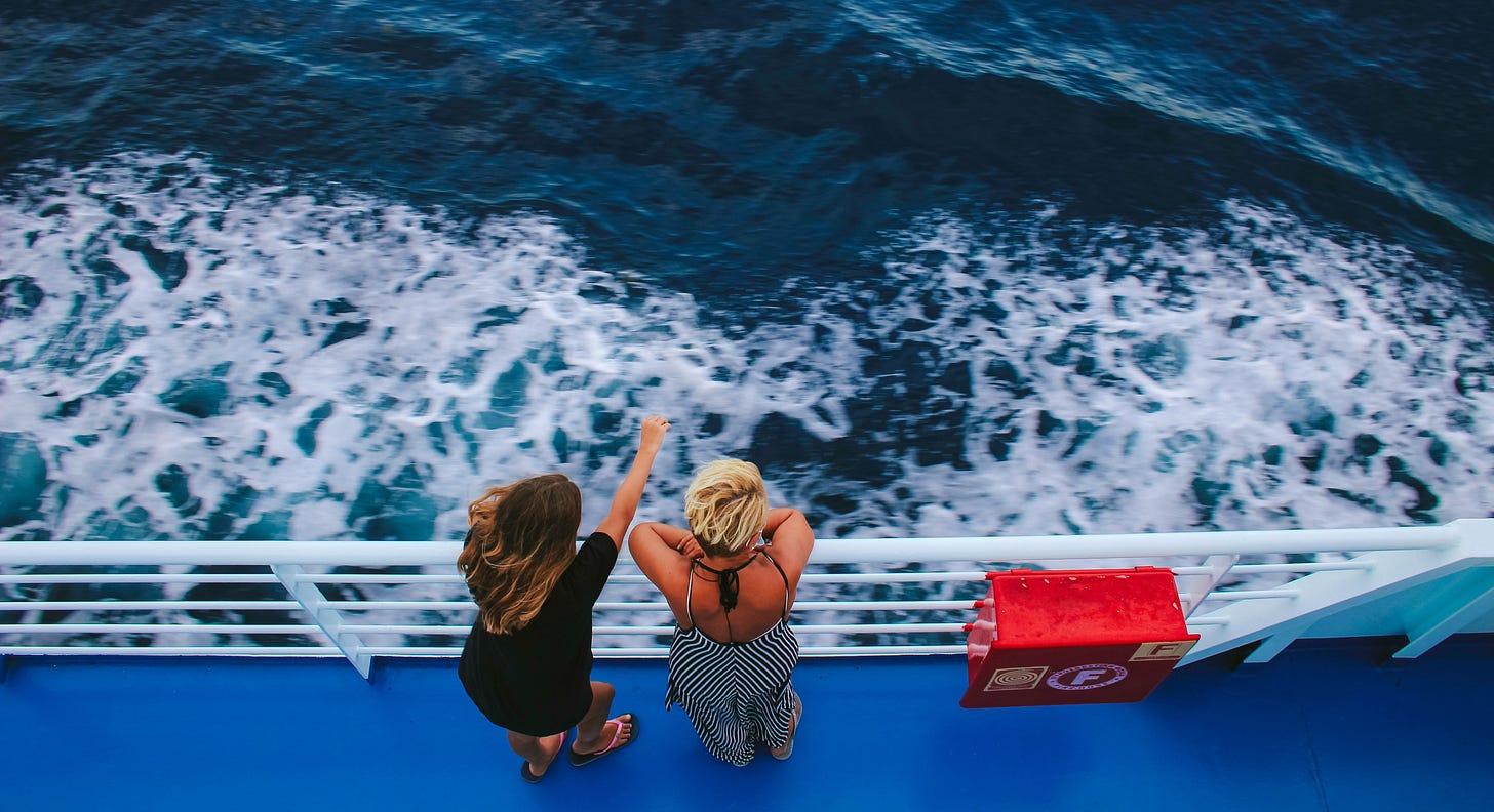 Two white women are standing at the deck of a cruise ship, looking out into the sea. Their backs are turned so you don't see their faces. 