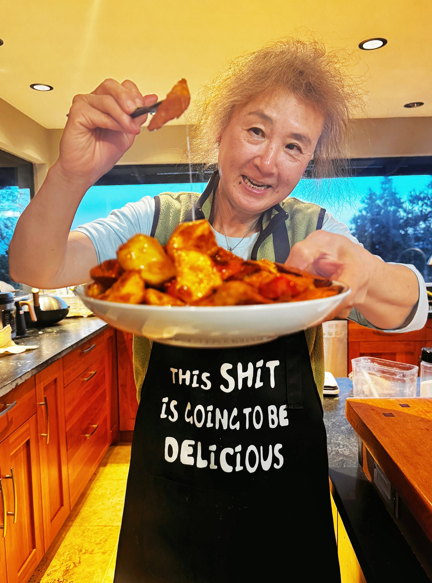 My mother holding a candied sweet potato dish with her left hand, and her right hand holing chopsticks with a piece of sweet potato. Her apron reads: This shit is going to be delicious.