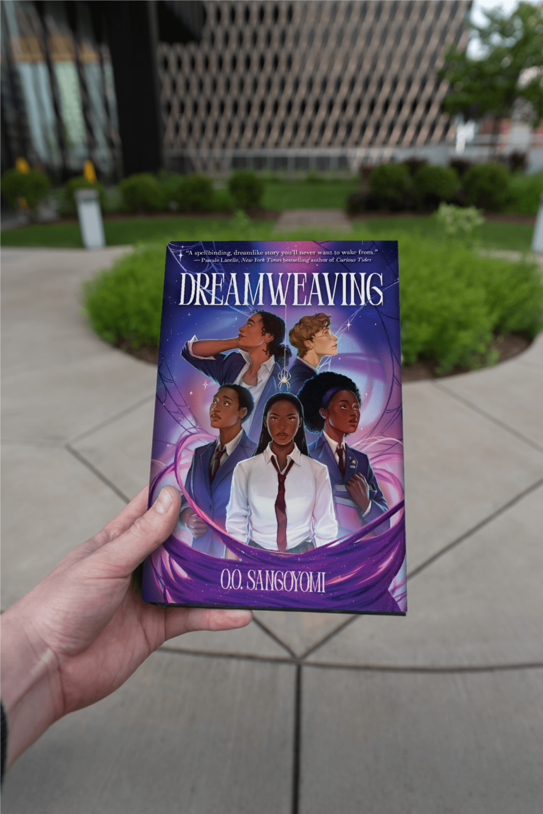 Book "Dreamweaving" held in front of a school building and park