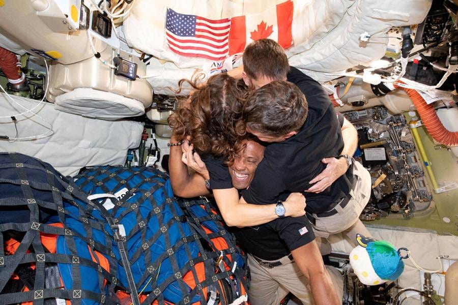 Four astronauts embrace inside a spacecraft and hug in front of American and Canadian flags. Four astronauts embrace inside a spacecraft and hug in front of American and Canadian flags.