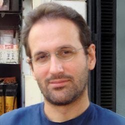 A imagem contém um homem com cabelo e barba escuro, ele usa óculos e camiseta azul