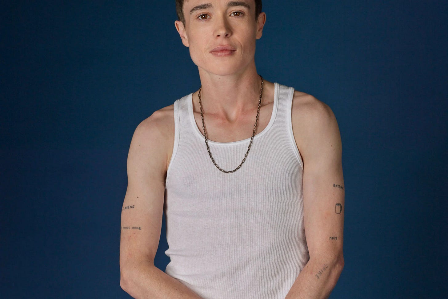 Elliot Page wearing a white tank and jeans, posing against blue background Elliot Page wearing a white tank and jeans, posing against blue background