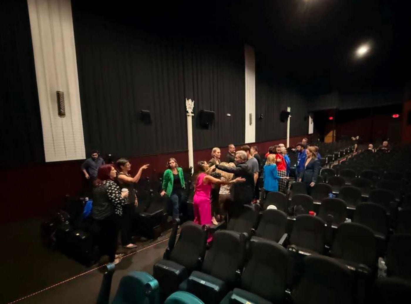 Speakers cluster in the front rows of an empty theater after dress rehearsal, several of them embracing, rows of empty seats sweeping away to the right. Speakers cluster in the front rows of an empty theater after dress rehearsal, several of them embracing, rows of empty seats sweeping away to the right.