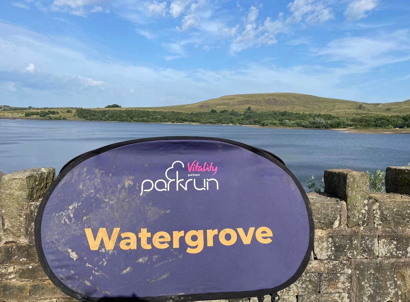 Watergrove parkrun sign in front of reservoir with low hills behind