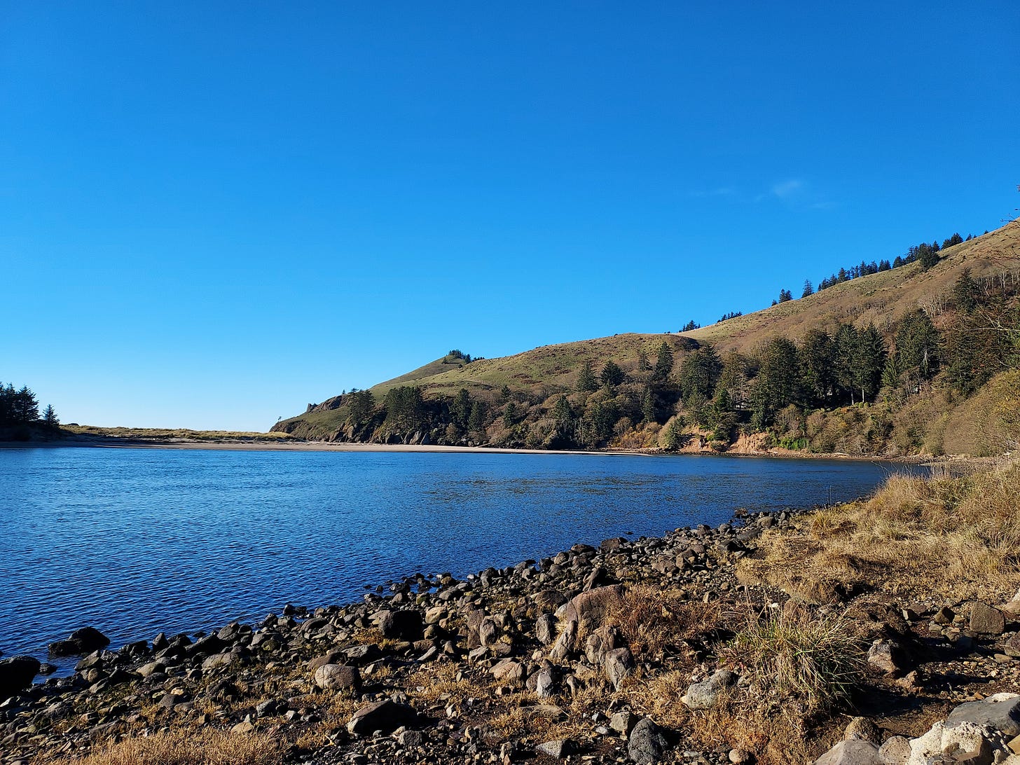 The top third of the picture is a robin's egg blue sky with no clouds. In the middle, a sloping brown and green spit of land terminates in a darker blue water, Foregrounded by rocks  casting long shadows in the afternoon sun.