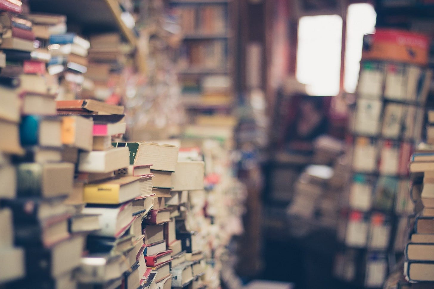 Image is of a room full of books with a window in the back. The books are on shelves, piled on tables, piled on the floor.