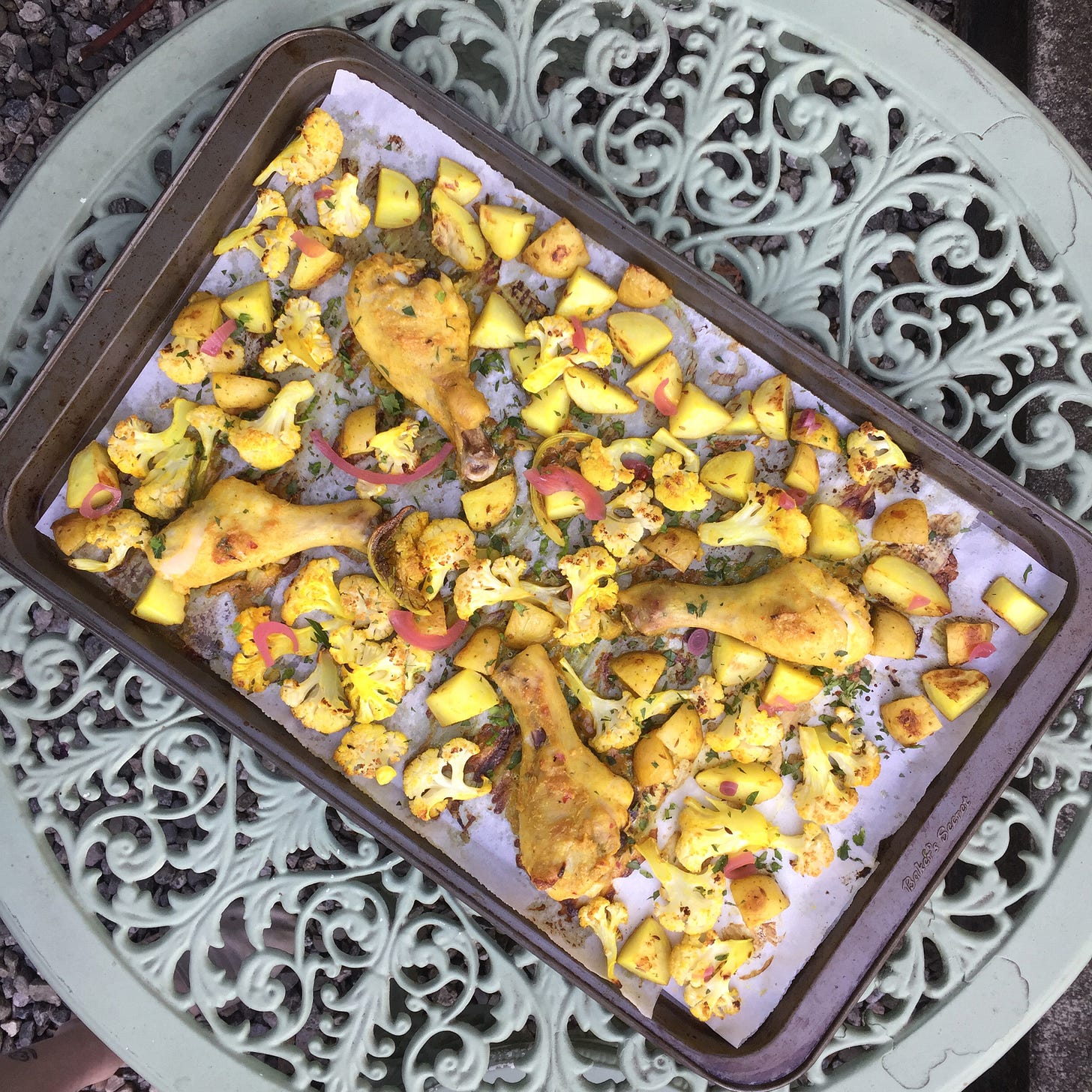 from above, a large sheet pan with four drumsticks in between roasted cauliflower and potatoes. Everything is tinged yellow from turmeric, and there are pickled red onions scattered over the dish.