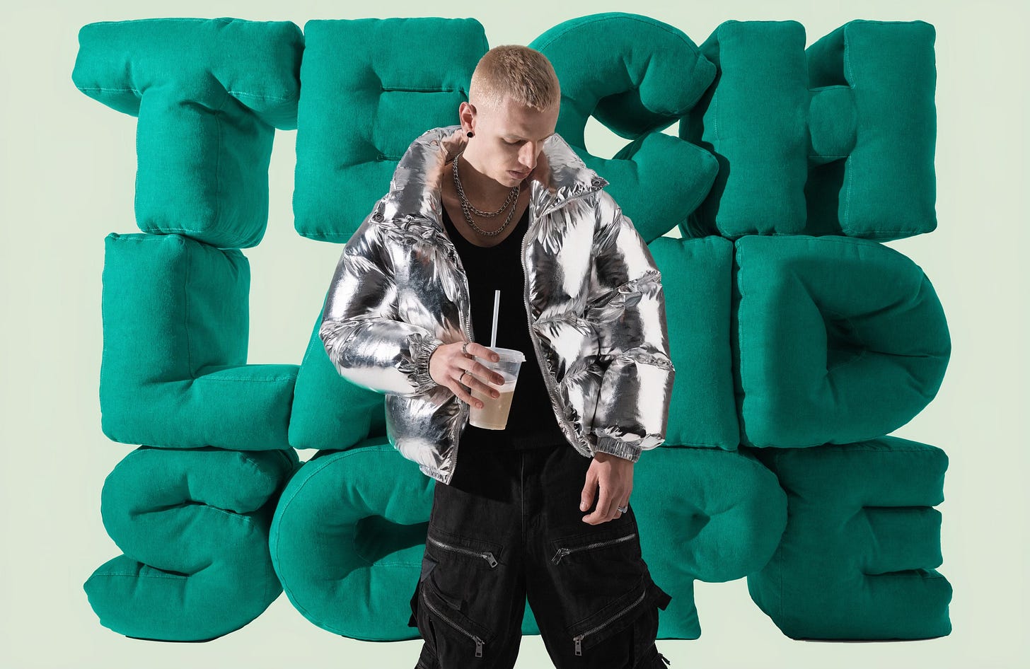 A young white British man, working class roadman style, skinny, short blonde hair, wearing a shiny silver puffer jacket and combat trousers, standing in a casual swaggering pose. Behind him are giant physical 3D foam letters stacked on top of each other, with the text "TECH LAND SCAPE" in huge slab sans-serif letters that fill the frame.