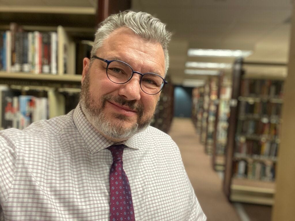 Photograph of a smiling middle-aged white man wearing glasses, a checkered button-down shirt and tie with library shelving in the background.