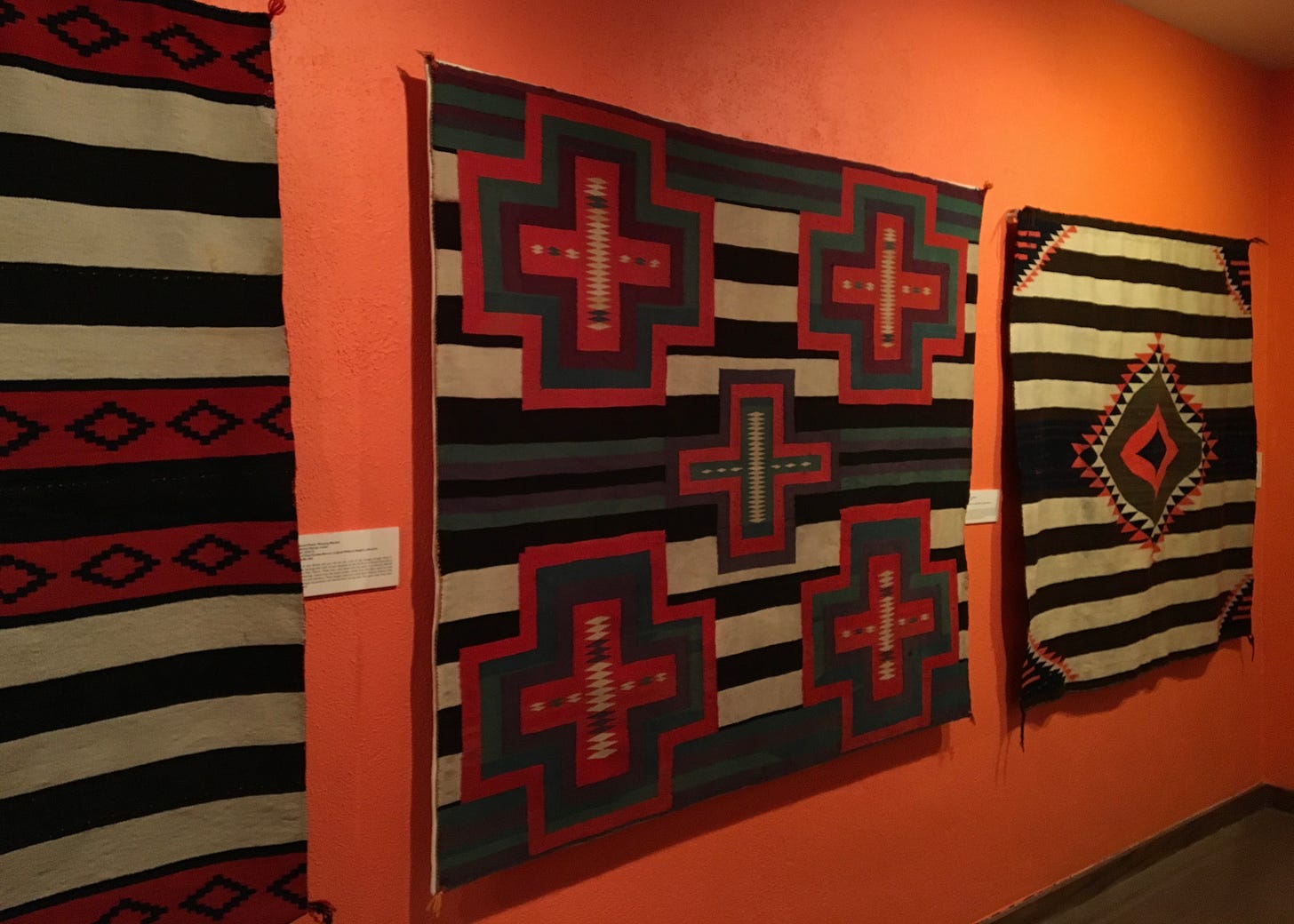 A historic Navajo wearing blanket displayed in a museum, featuring horizontal stripes and cross motifs in red, dark, and natural tones. Collection of the Millicent Rogers Museum, Taos, New Mexico. A historic Navajo wearing blanket displayed in a museum, featuring horizontal stripes and cross motifs in red, dark, and natural tones. Collection of the Millicent Rogers Museum, Taos, New Mexico.