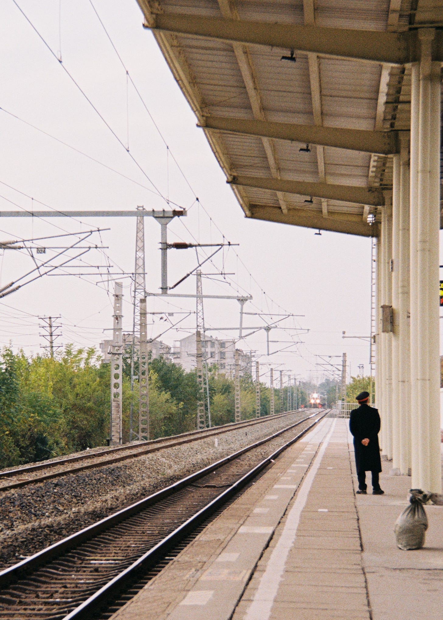 A railway photograph with strong perspective. On the right, a railway staff member in a dark long coat and peaked cap stands on the platform of Yingcheng Zhan (Yingcheng Station), looking down the tracks. On the left, railway tracks and complex overhead lines stretch into the distance. At the far end of the tracks, the faint headlights of an approaching locomotive are visible. The image has a vintage, film-like ae