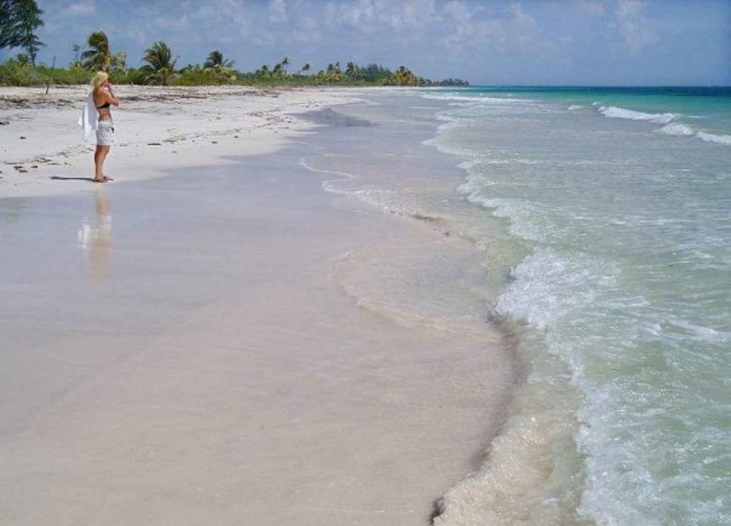 A woman standing alone at the shoreline of a wide, empty white-sand Caribbean beach lined with palms. A woman standing alone at the shoreline of a wide, empty white-sand Caribbean beach lined with palms.
