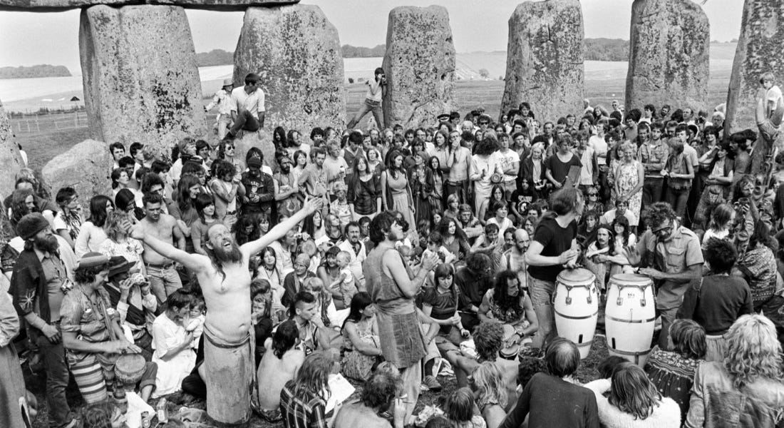 A black and white picture of a group of people crowded around a man with his arms outstretched, at Stonehenge
