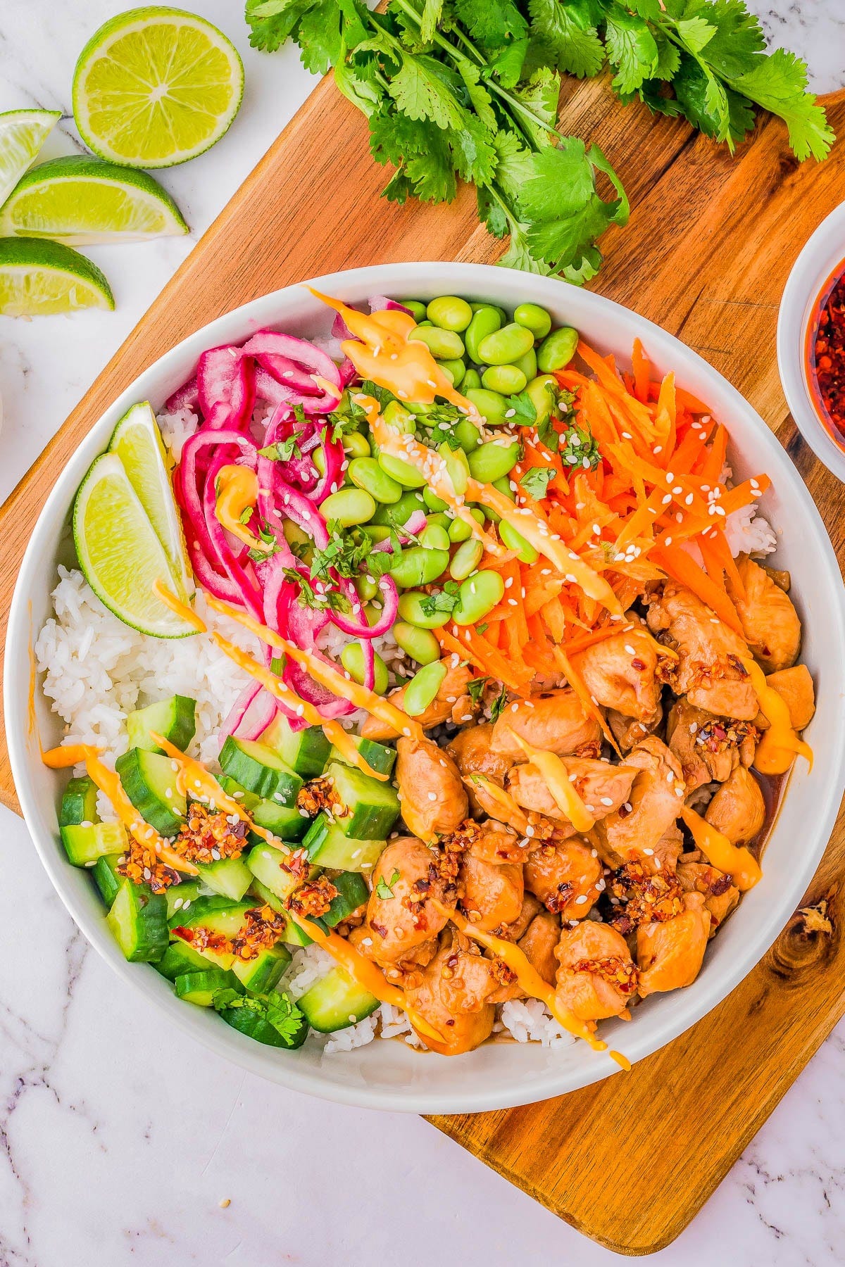 A colorful poke bowl with chicken, rice, edamame, cucumber, carrots, pickled onions, cilantro, lime, and sesame seeds, served on a wooden board. A colorful poke bowl with chicken, rice, edamame, cucumber, carrots, pickled onions, cilantro, lime, and sesame seeds, served on a wooden board.